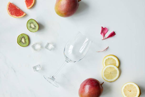 flat lay with empty wineglass, flower petals, ice cubes and assorted exotic fruits on white tabletop - Zdjęcie, obraz