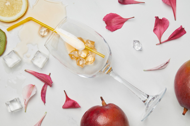 top view of arrangement of exotic fruits, flower petals, wineglass with straw and ice cubes on white surface - Photo, image
