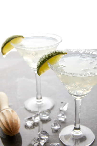 Selective focus of alcohol margarita cocktails with lime pieces and wooden squeezer on grey surface on white - Foto, afbeelding