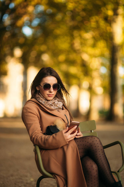 Una giovane ragazza con un cappotto marrone si siede in mezzo al parco su uno sgabello kaki
 - Foto, immagini
