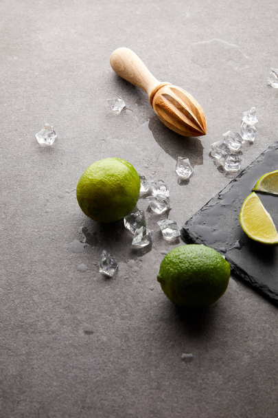 close up view of wooden squeezer, limes and ice cubes for cocktail on grey surface - Zdjęcie, obraz