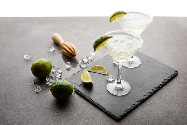 close up view of alcohol cocktails with pieces of lime, ice cubes and wooden squeezer on grey tabletop - Foto, imagen