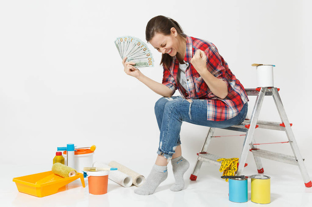 Женщина держит связку долларов, наличные деньги делают жест победителя, сидя на лестнице с инструментами для ремонта квартиры изолированы на белом фоне. Обои, инструменты для рисования. Ремонт дома концепции
 - Фото, изображение