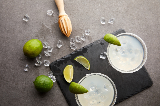 Top view of margarita cocktails with pieces of lime, ice cubes and wooden squeezer on grey tabletop - Foto, Bild