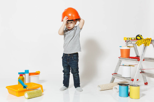 Little boy in orange protective helmet with instruments for renovation apartment isolated on white background. Wallpaper, gluing accessories, painting tools. Repair home. Parenthood, childhood concept - Valokuva, kuva