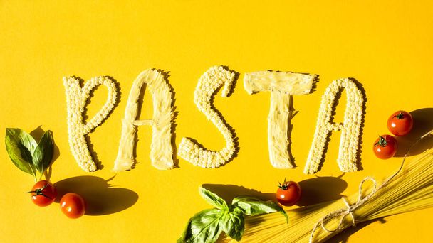 A word pasta is laid out of small pasta on a yellow background next to cherry tomatoes and a leaf of basil. Ingredients for a ready-made dish of spaghetti. Top view, flat lay - 写真・画像