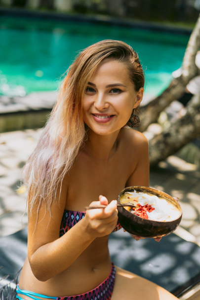 beautiful young woman in bikini eating tropical dessert and smiling at camera - Foto, Bild