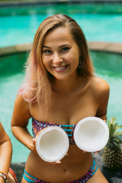 beautiful cheerful girl in bikini holding halved coconut and smiling at camera - Foto, immagini