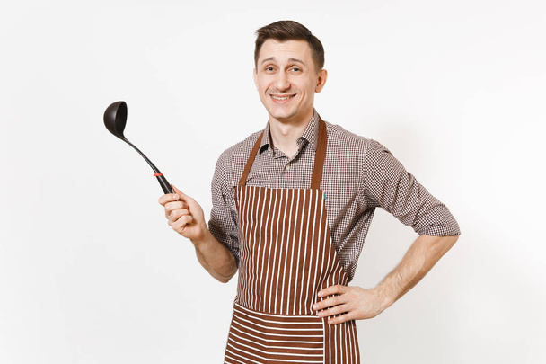 Young smiling man chef or waiter in striped brown apron, shirt holding black ladle or kitchen spoon isolated on white background. Male housekeeper or houseworker. Kitchenware and cuisine concept. - Photo, Image
