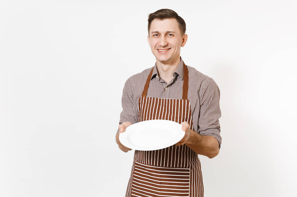 Nuori mies kokki tai tarjoilija raidallinen ruskea esiliina, paita tilalla valkoinen pyöreä tyhjä kirkas levy eristetty valkoisella pohjalla. Miestaloudenhoitaja tai taloudenhoitaja. Kotityöntekijän kopiotila mainontaa varten
 - Valokuva, kuva