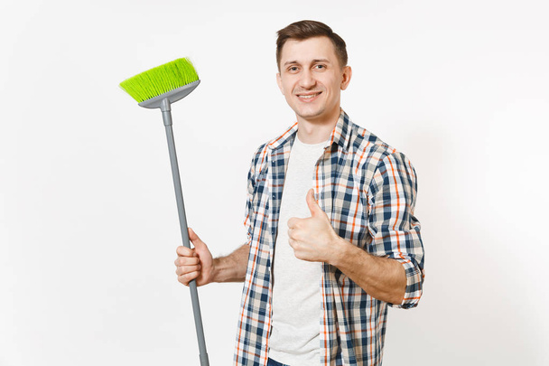 若い笑顔幸せな家政婦男チェッカー シャツ ホワイト バック グラウンド上に分離されて緑のほうきで掃除を押しながら。家事をしている男性。広告のためのスペースにコピーします。清浄度の概念. - 写真・画像