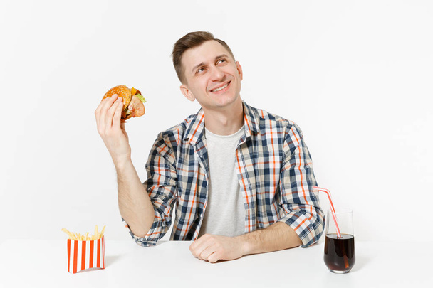 Uśmiechający się młody człowiek w koszuli siedzi przy stole z burger, frytki, cola szklanki na białym tle. Prawidłowe odżywianie lub klasyczny amerykański fast food. Reklama obszar z miejsca kopii. - Zdjęcie, obraz
