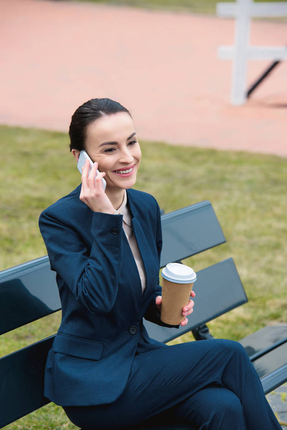 mosolygó üzletasszony, beszél a smartphone és a gazdaság kávé papírpohár padon - Fotó, kép
