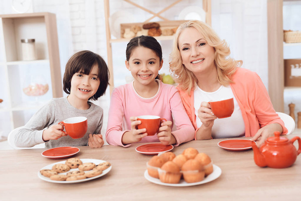 Молодая бабушка с внуком и внучкой едят печенье и пьют чай в красных кружках на кухне
 - Фото, изображение