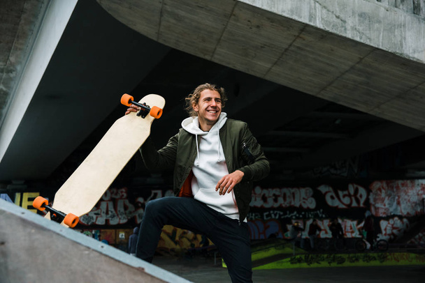 Longboard es su pasión. Guapo joven longboarding y mirando hacia otro lado con sonrisa en el parque de skate al aire libre
  - Foto, imagen