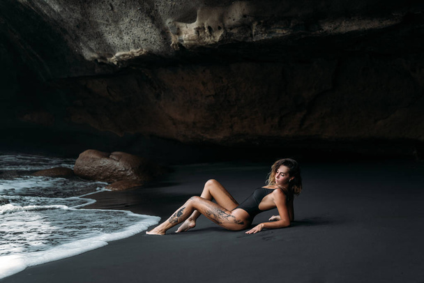 Beautiful girl with sexy body in black swimsuit lying on the beach with black volcanic sand. Young model with curly hairs, tanned skin posing and relaxing in the journey. - Valokuva, kuva