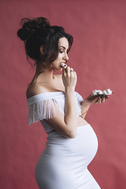 Beautiful pregnant model with long brown hair in the white dress smiles and eats marshmallows, portrait of beautiful pregnant woman, cute pregnant belly, future mother - Photo, Image