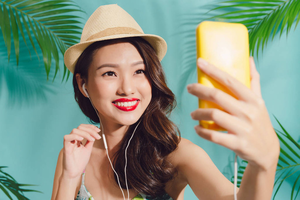 Pretty smiling asian woman using smartphone taking selfie standing over blue background - Zdjęcie, obraz