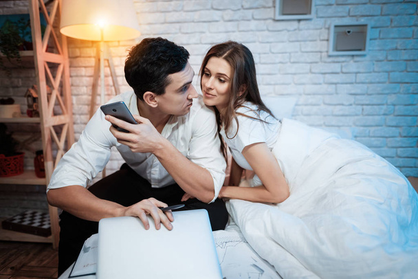 businessman holding smartphone while wife distracting husband from work in bedroom - Foto, Imagem