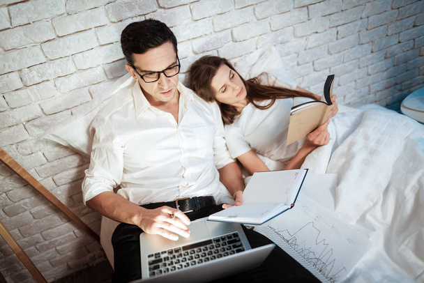 Young businessman working on laptop while woman reading book in bed - Photo, Image