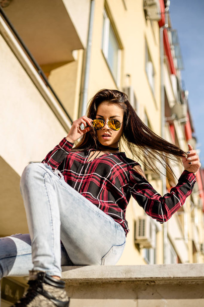 beautiful brunette woman in sunglasses sitting on parapet at sunny day   - Foto, Imagen