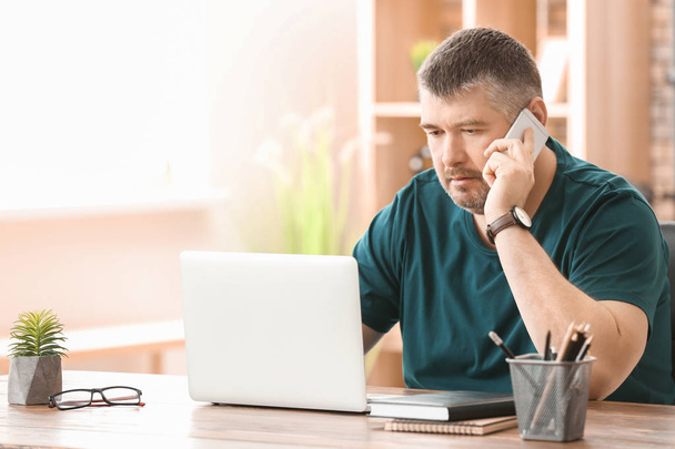 Mature man talking on phone while working with laptop indoors - Foto, immagini
