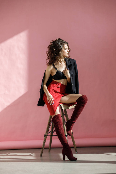 Fashion studio portrait of beautiful curly woman wearing red skirt, black jacket and black lingerie, sitting on wooden chair over pink background - Фото, зображення
