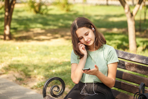 beautiful teenage girl sitting on benches in park, in hands with phone and wearing headphones. - Photo, image