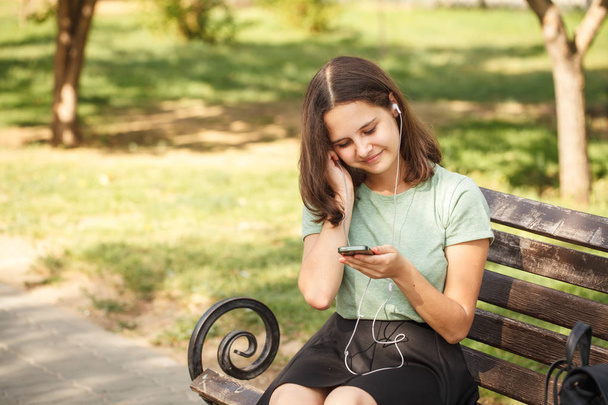 beautiful teenage girl sitting on benches in park, in hands with phone and wearing headphones. - Фото, изображение