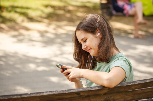 beautiful teenage girl sitting on benches in park, in hands with phone. Close-up. - Zdjęcie, obraz