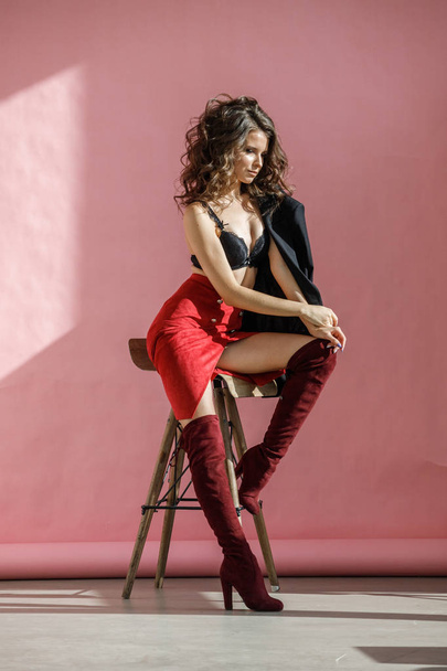 Fashion studio portrait of beautiful curly woman wearing red skirt, black jacket and black lingerie, sitting on wooden chair over pink background - Photo, image