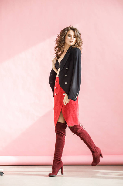 Fashion studio portrait of beautiful curly woman wearing red skirt, black jacket and black lingerie, standing over pink background - Fotografie, Obrázek