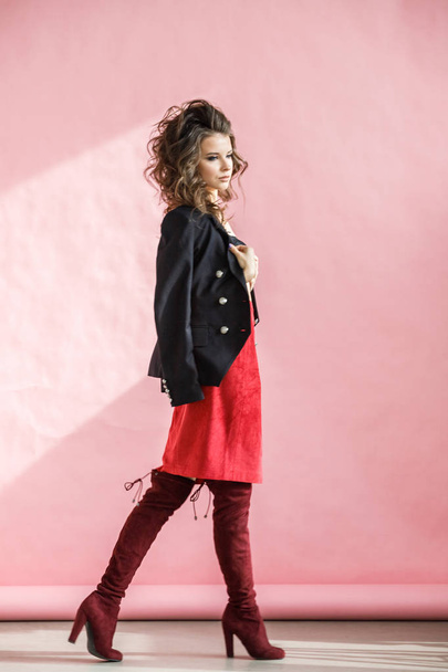 Fashion studio portrait of beautiful curly woman wearing red skirt, black jacket and black lingerie, standing over pink background - Foto, afbeelding