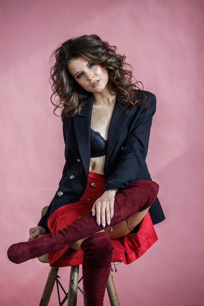 Fashion studio portrait of beautiful curly woman wearing red skirt, black jacket and black lingerie, sitting on wooden chair over pink background - Fotó, kép