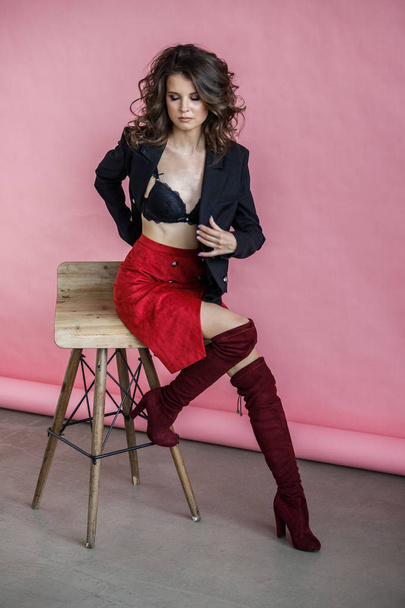Fashion studio portrait of beautiful curly woman wearing red skirt, black jacket and black lingerie, sitting on wooden chair over pink background - Foto, Imagen