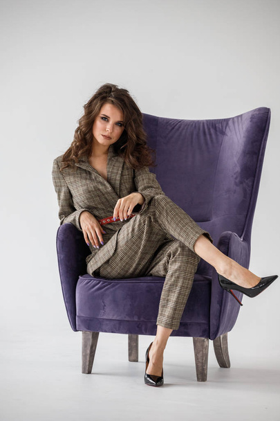 Fashion studio portrait of beautiful curly woman wearing checkered casual suit with red belt, sitting in purple armchair, white wall background  - Foto, Bild