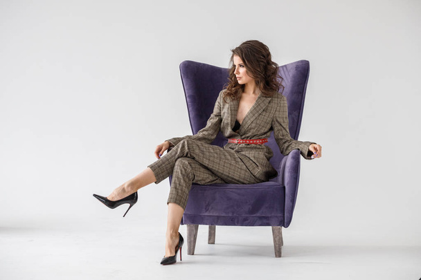 Fashion studio portrait of beautiful curly woman wearing checkered casual suit with red belt, sitting in purple armchair, white wall background  - Fotó, kép