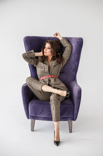 Fashion studio portrait of beautiful curly woman wearing checkered casual suit with red belt, sitting in purple armchair, white wall background  - Foto, Bild