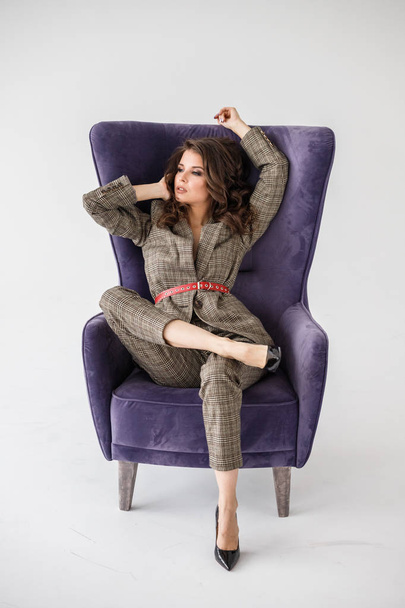 Fashion studio portrait of beautiful curly woman wearing checkered casual suit with red belt, sitting in purple armchair, white wall background  - Valokuva, kuva