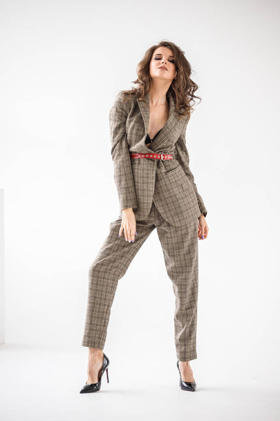 Fashion studio portrait of beautiful curly woman wearing checkered casual suit with red belt, standing on white wall background  - Foto, immagini