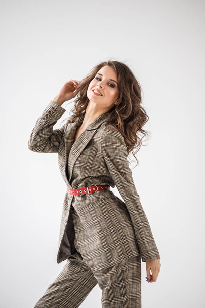 Fashion studio portrait of beautiful curly woman wearing checkered casual suit with red belt, standing on white wall background  - Zdjęcie, obraz