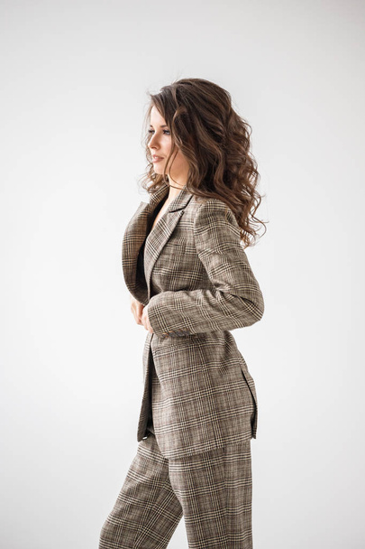Fashion studio portrait of beautiful curly woman wearing checkered casual suit and black bra, standing on grey wall background with red belt in hands - Fotó, kép
