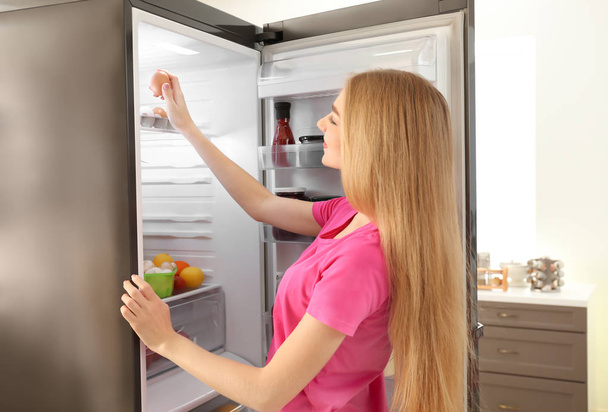 Young woman taking egg from refrigerator in kitchen - Foto, afbeelding
