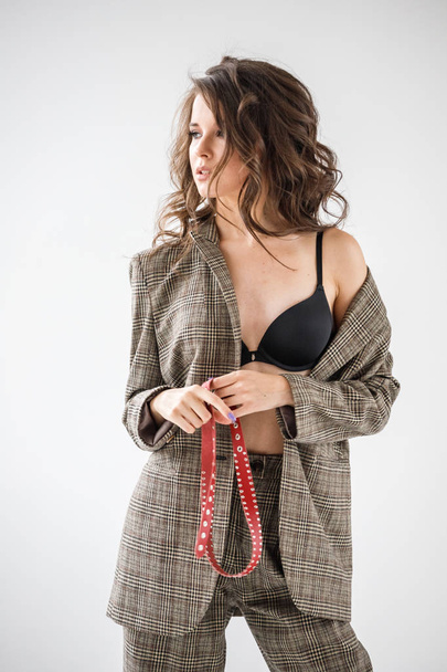 Fashion studio portrait of beautiful curly woman wearing checkered casual suit and black bra, standing on grey wall background with red belt in hands - Photo, image