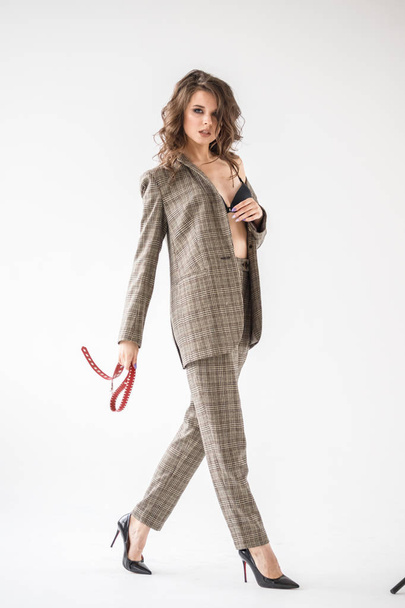 Fashion studio portrait of beautiful curly woman wearing checkered casual suit and black bra, standing on grey wall background with red belt in hands - 写真・画像