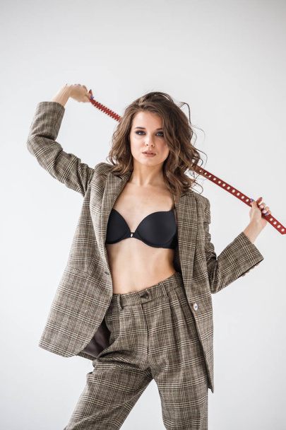Fashion studio portrait of beautiful curly woman wearing checkered casual suit and black bra, standing on grey wall background with red belt in hands - Φωτογραφία, εικόνα