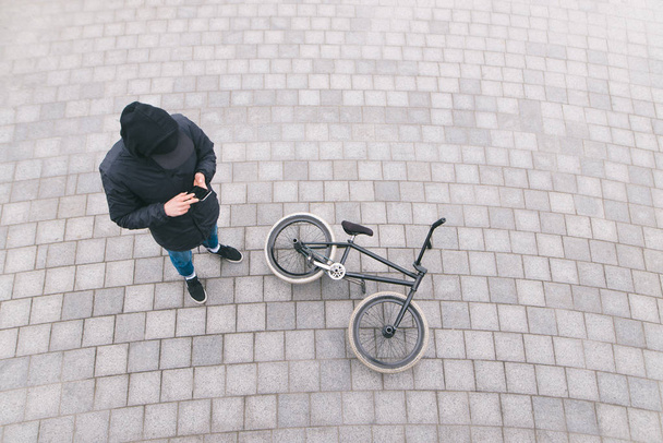 Ένας νεαρός άνδρας στέκεται στον δρόμο κοντά ένα ποδήλατο Bmx και χρησιμοποιεί ένα smartphone. Ξεκούραση μετά από κόλπα για Bmx. Top view - Φωτογραφία, εικόνα