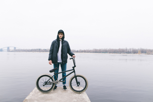 Портрет всадника BMX, стоящего с велосипедом на фоне реки. Прогулка с BMX на велосипеде. BMX
 - Фото, изображение