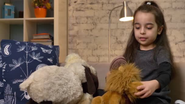 A little girl sitting on the couch and playing with a teddy bear and a lion, combs the fur of a toy lion, home comfort in the background 50 fps - Filmati, video