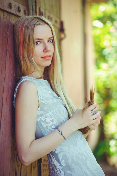 Přitažlivá skromná Mladá blondýnka na červeném pozadí dřevěná vlasy jí maloval v růžové pruhované pruhované, v modrých šatech plachý a zvídavý - Fotografie, Obrázek